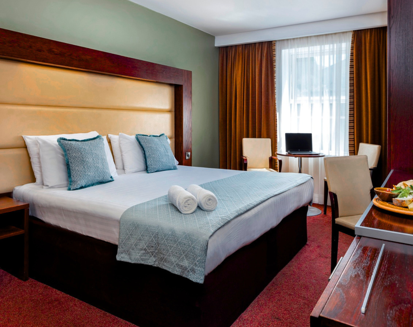 Stylish hotel room with a cosy bed, modern desk, and elegant dining area featuring a tempting fruit platter and wine.