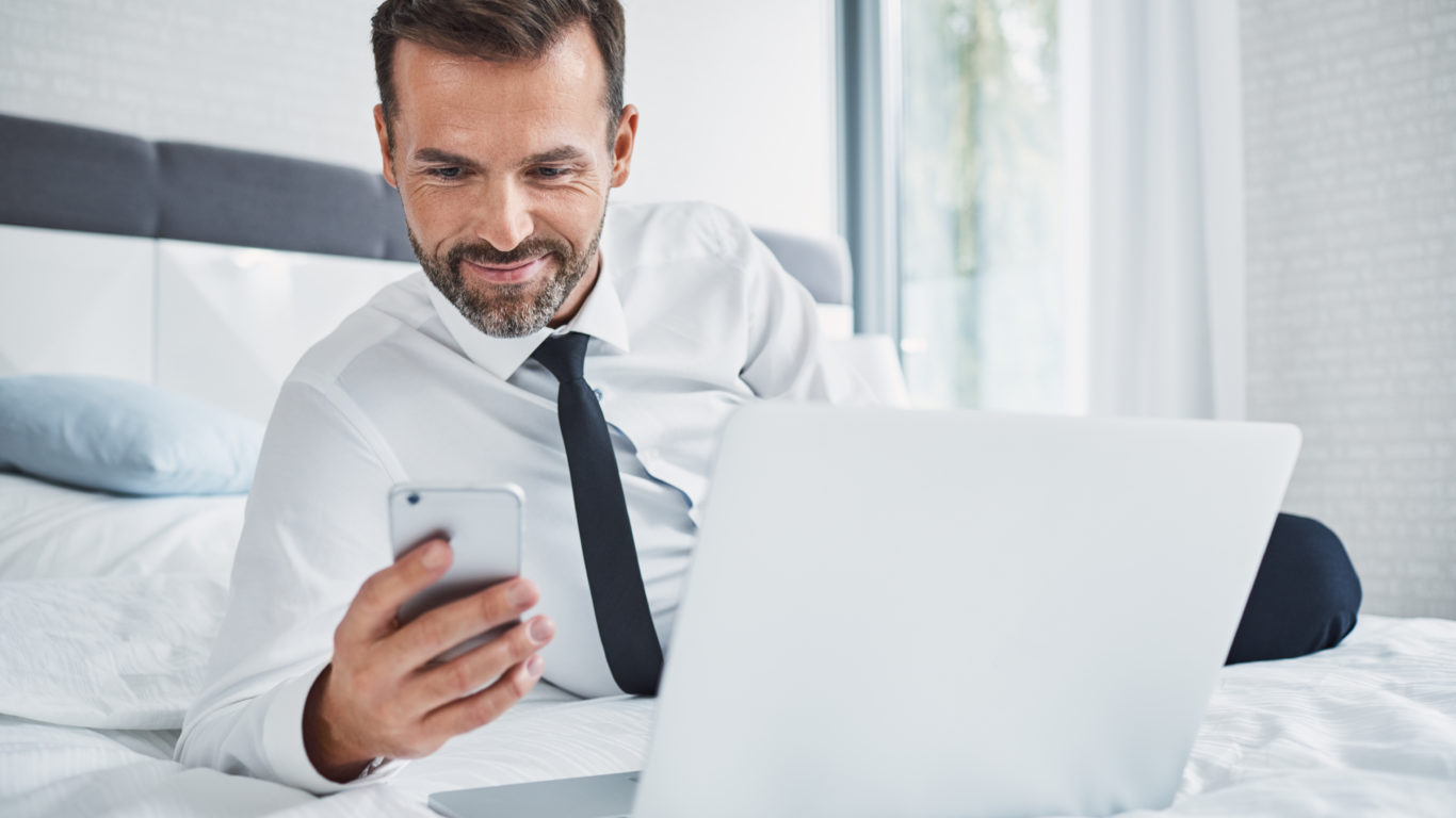 Smiling businessman using phone while lying on bed in hotel room on business trip