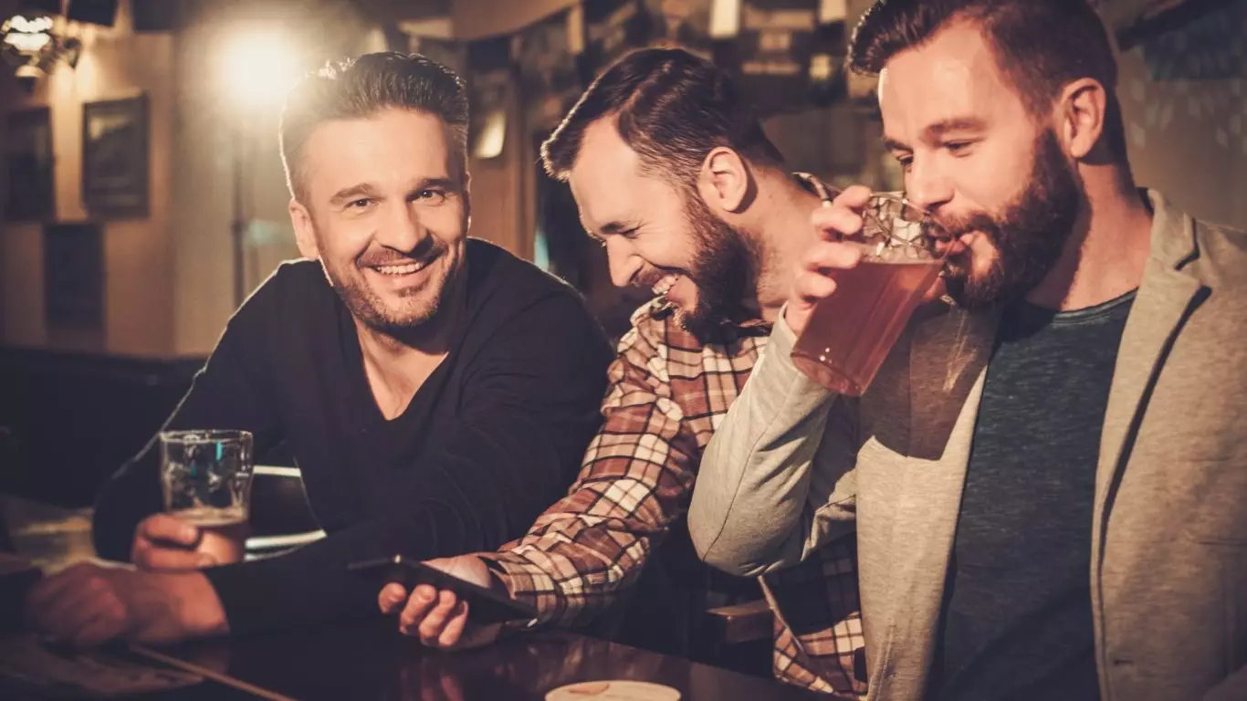 Cheerful old friends having fun with smartphone and drinking draft beer at bar counter in pub.