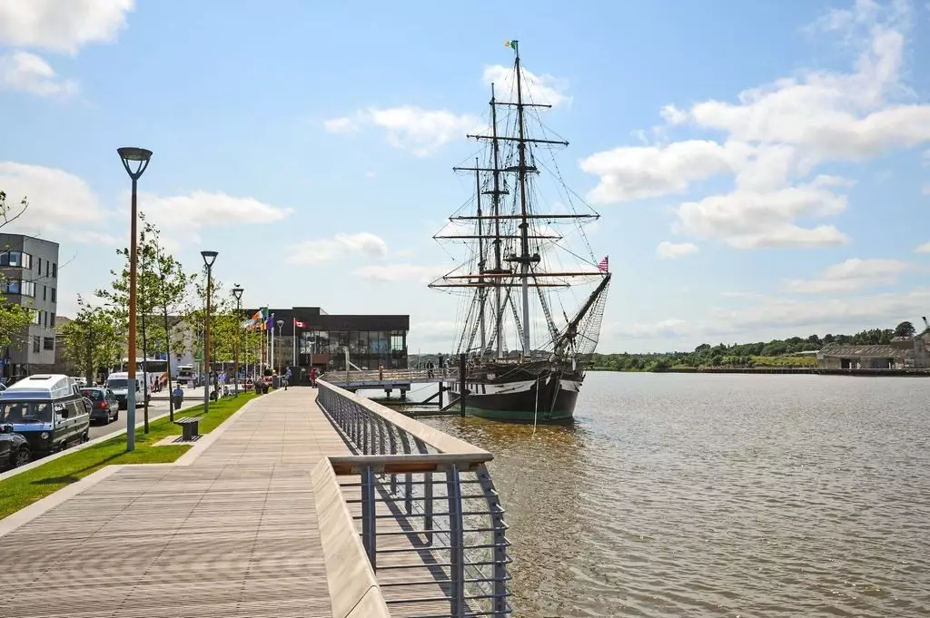 Scenic riverside view with a classic sailing ship, modern buildings, lush trees, and clear blue sky, perfect for exploring.