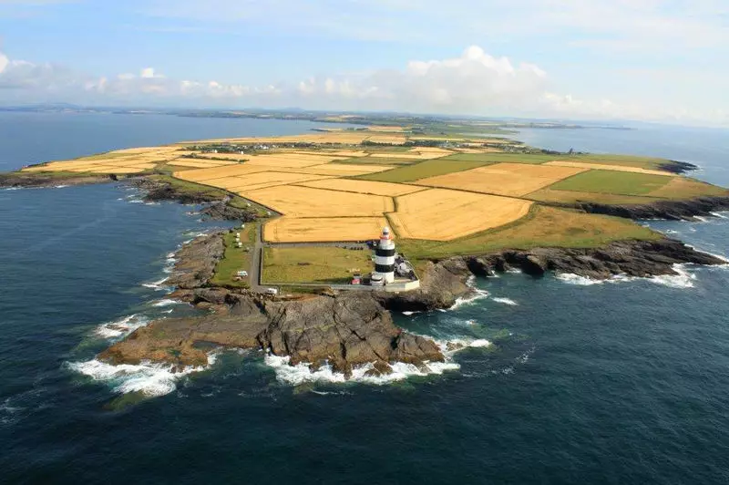 Aerial view of stunning lighthouse on rocky Irish coastline, surrounded by golden fields under a clear blue sky.