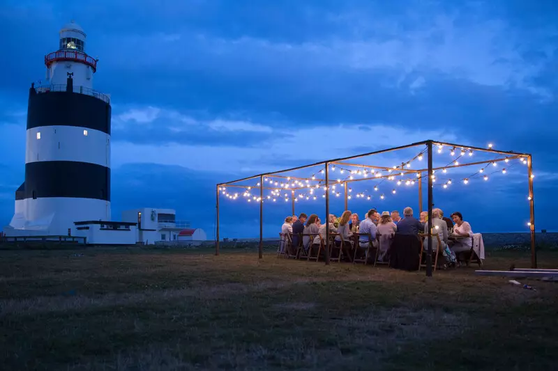 Guests enjoy candlelit dinner under twinkling lights by historic lighthouse, creating a magical seaside dining experience.