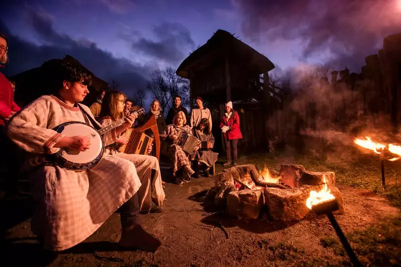 Guests enjoy live folk music by a warm campfire at dusk, surrounded by rustic cabins and a tranquil wooded setting.