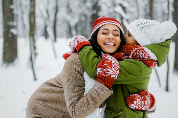 Happy couple in winter clothes laughing and hugging in a snowy forest, surrounded by snow-covered trees.