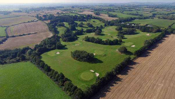 Aerial view of a golf course surrounded by fields and trees, with visible sand bunkers and fairways under a clear sky.