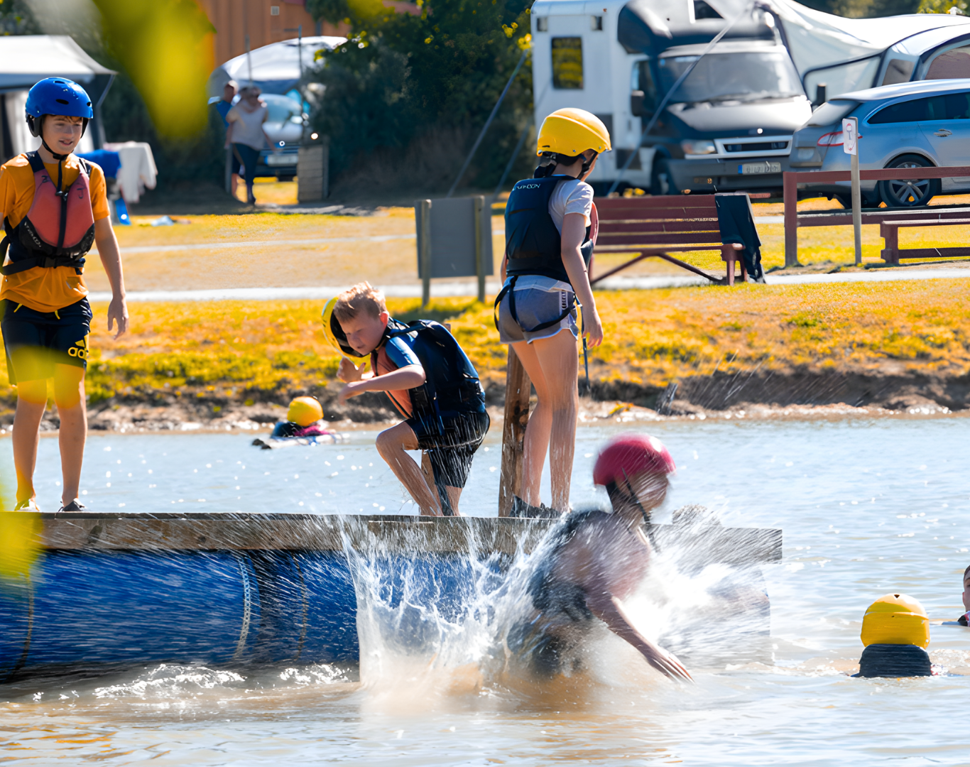 Families enjoy water activities with helmets and life vests at lakeside resort, with caravans in the background.