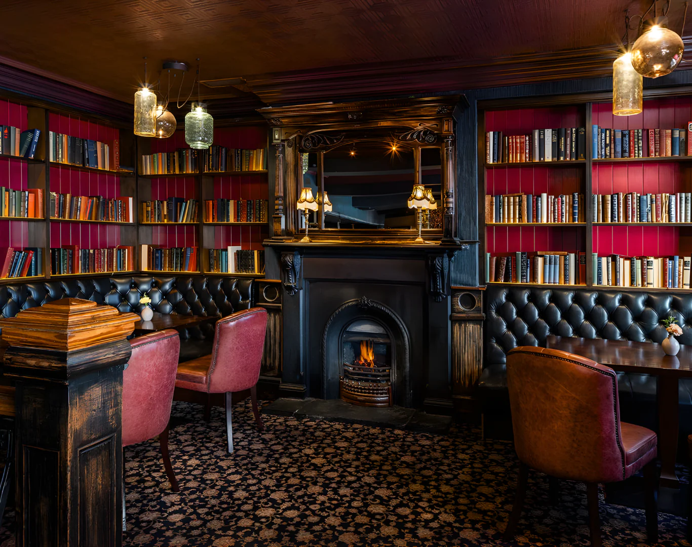 Cosy hotel library lounge with plush seating, warm fireplace, and elegant red bookshelves for a relaxing experience.