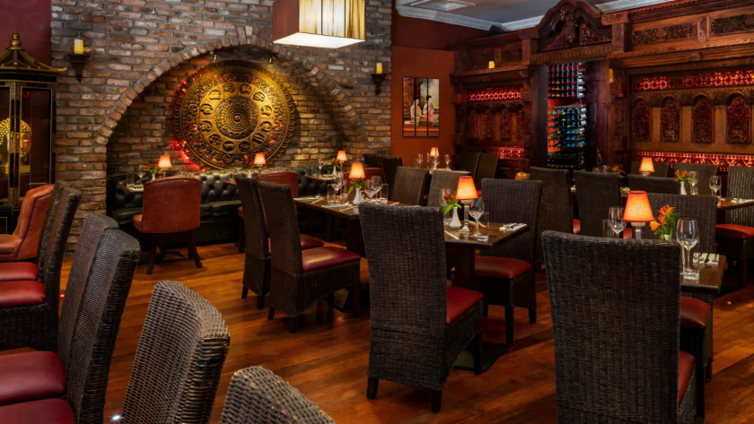 Cosy hotel dining room with elegant wicker chairs, ornate wall design, warm lighting, and richly decorated woodwork.