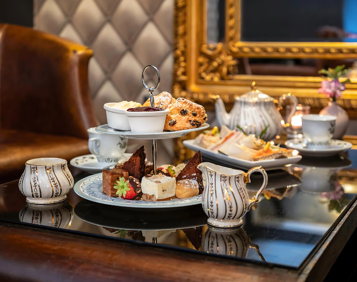 Elegant afternoon tea with scones, cakes, and sandwiches in a cosy hotel lounge with plush seating and ornate mirrors.