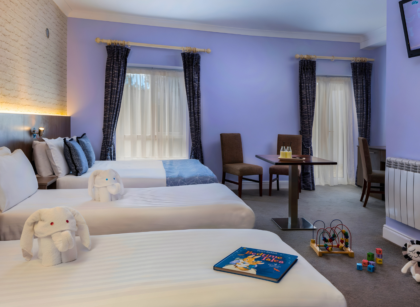 Family-friendly hotel room with three white beds, playful soft toys, children's book, and a wall-mounted flat-screen TV.